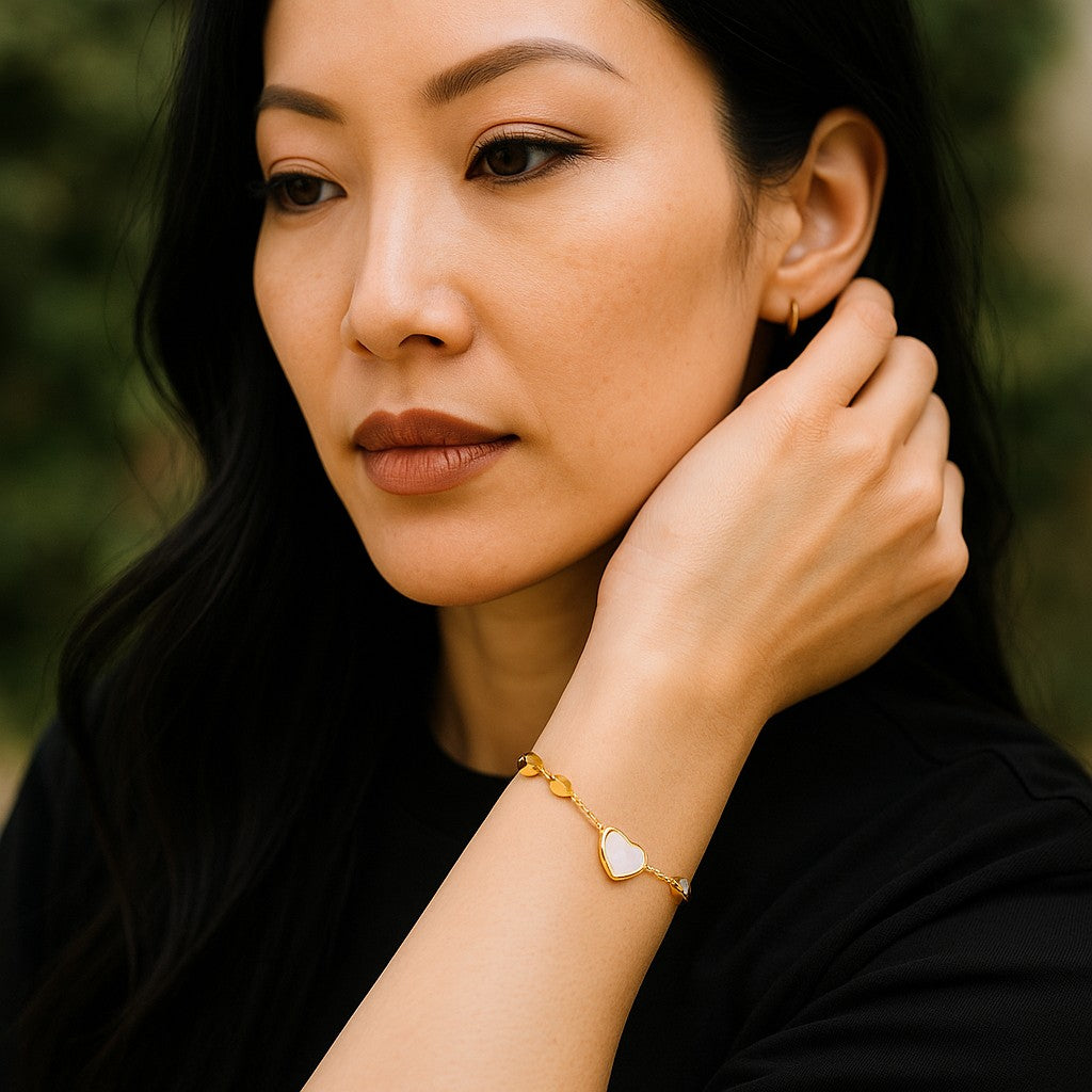 Woman wearing a gold bracelet with a heart charm, adjusting an earring against a blurred green background.